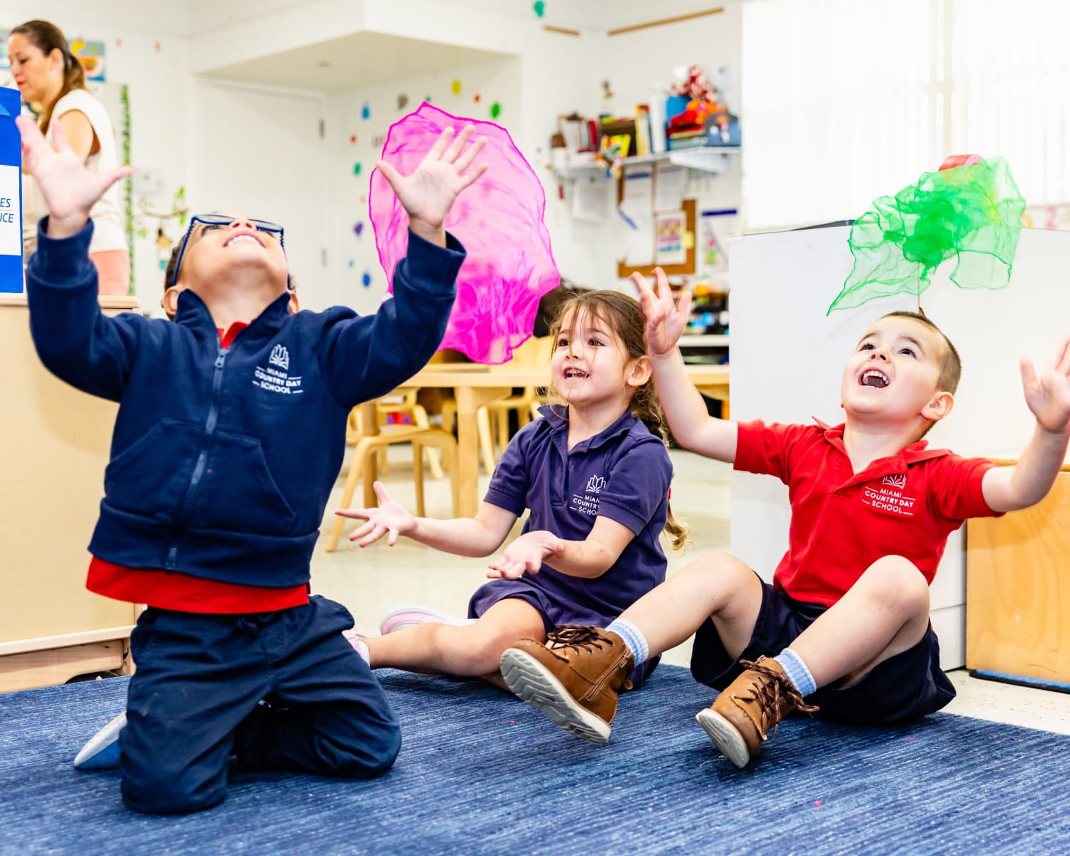 Young students in school uniforms sitting on a classroom rug joyfully tossing colorful scarves in the air