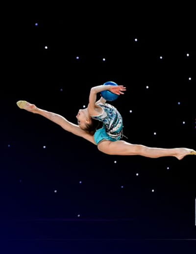Rhythmic gymnast in a blue leotard leaping mid-air while holding a ball during a performance