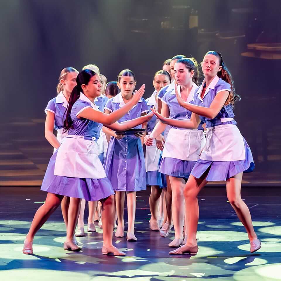 Group of young performers in purple waitress-style costumes dancing together on stage
