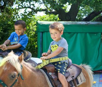 Two young boys smiling while riding a pony together at summer camp