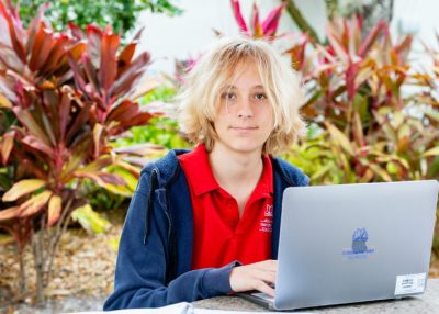 Student in a red school polo shirt sitting outdoors with a laptop, surrounded by tropical plants