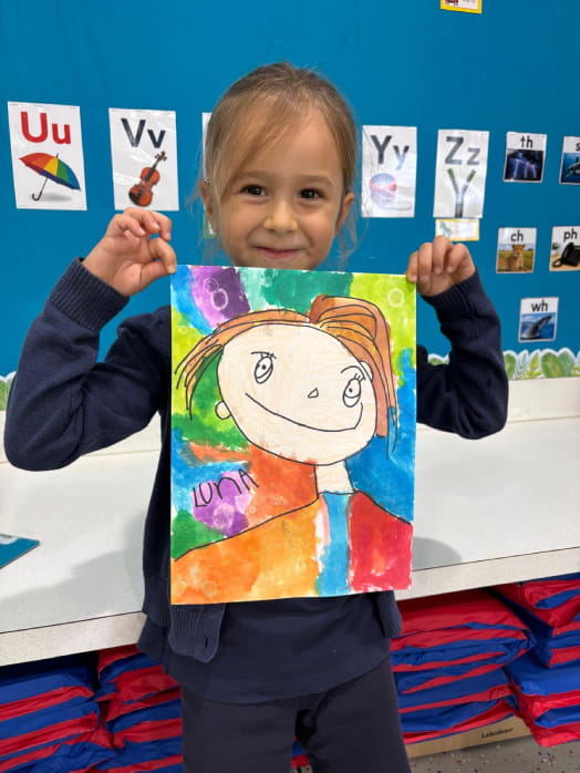 Junior kindergarten student smiling while holding up her colorful self-portrait artwork