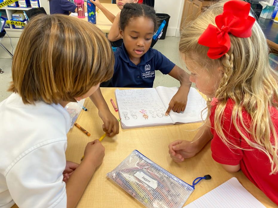 Three pre-k students in school uniforms working together on a writing activity at their desk